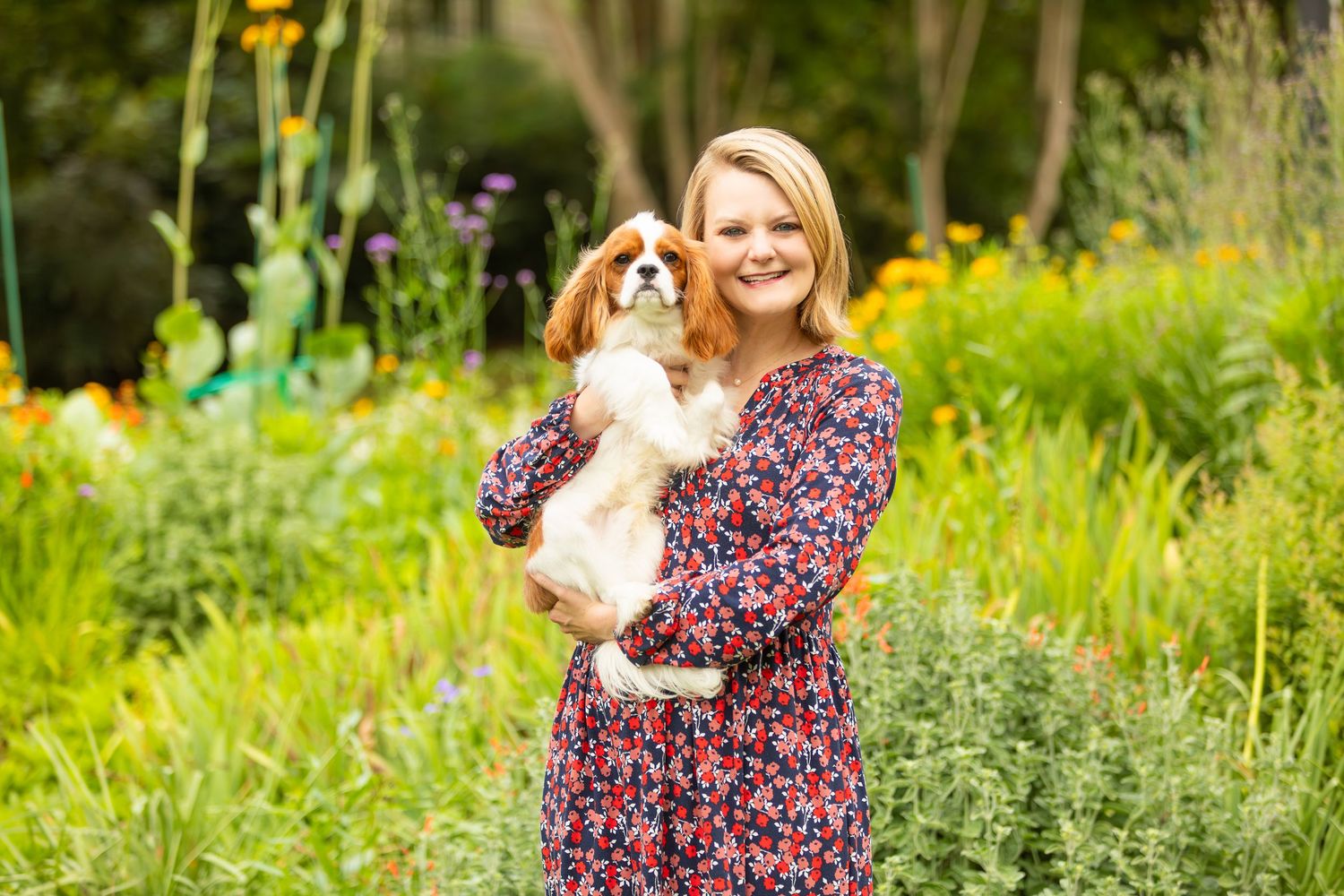 Someone in a floral dress holds a happy Cavalier King Charles Spaniel dog in a sunny garden setting.
