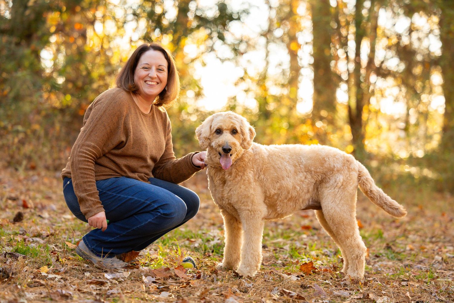 Woman in brown sweater crouching beside a golden doodle dog in a sunlit autumn forest setting.