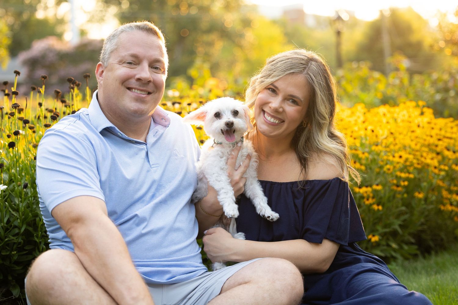 Smiling couple sitting outdoors in a garden holding a small white dog, surrounded by yellow flowers in warm sunlight.