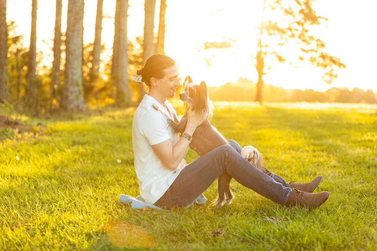 A figure sitting on sunny grass with their dog during golden hour sunset with trees in the background.