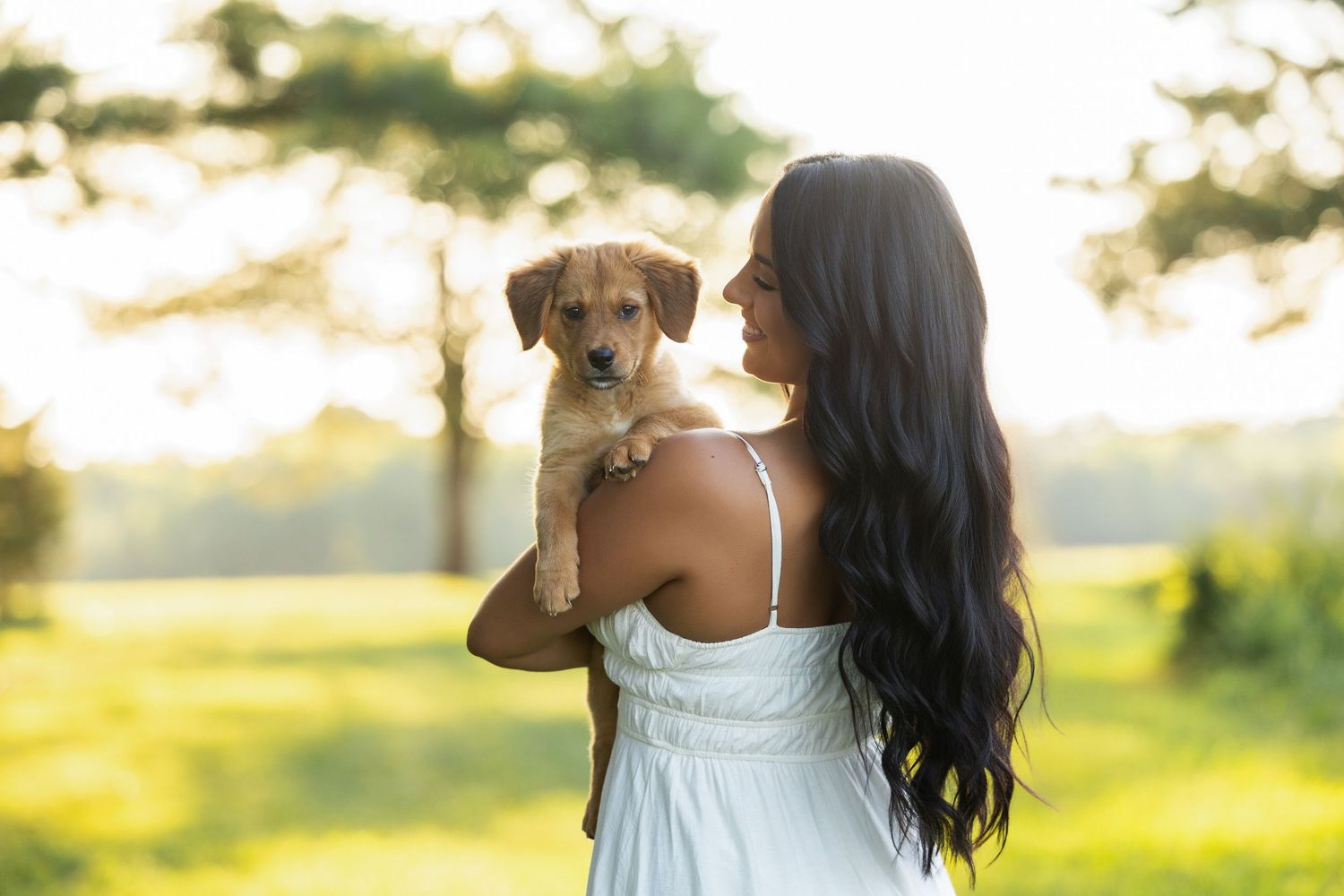 Woman in white dress holding a golden puppy outdoors in sunlit green park setting.