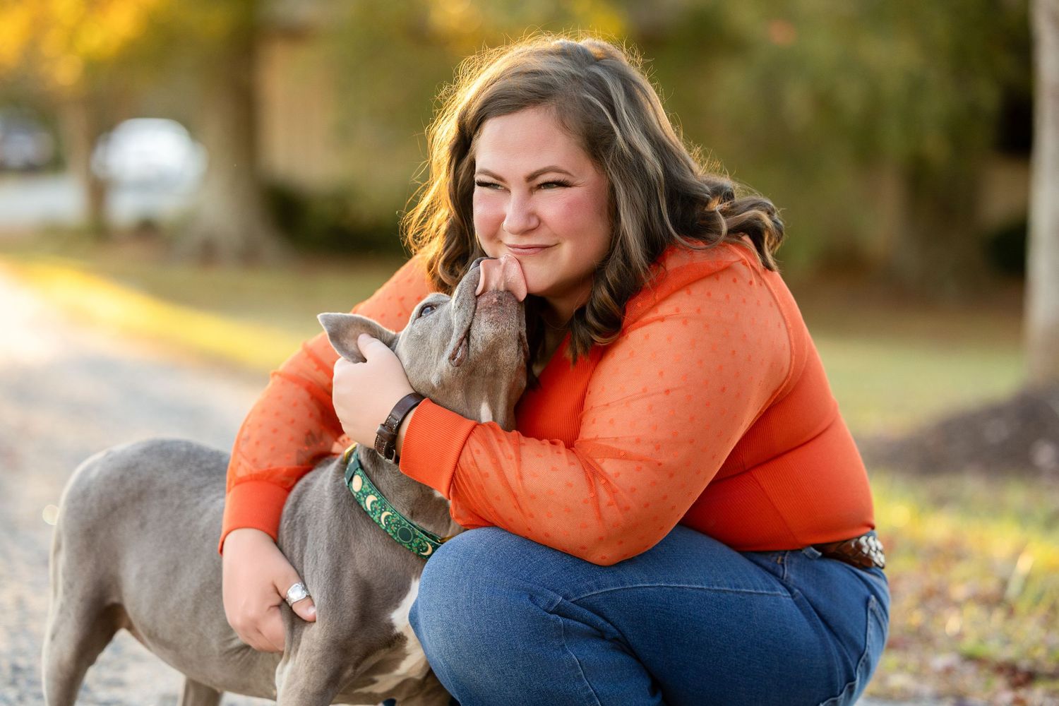 Person in orange sweater crouches down to embrace and pet a Weimaraner dog during golden hour outdoors.