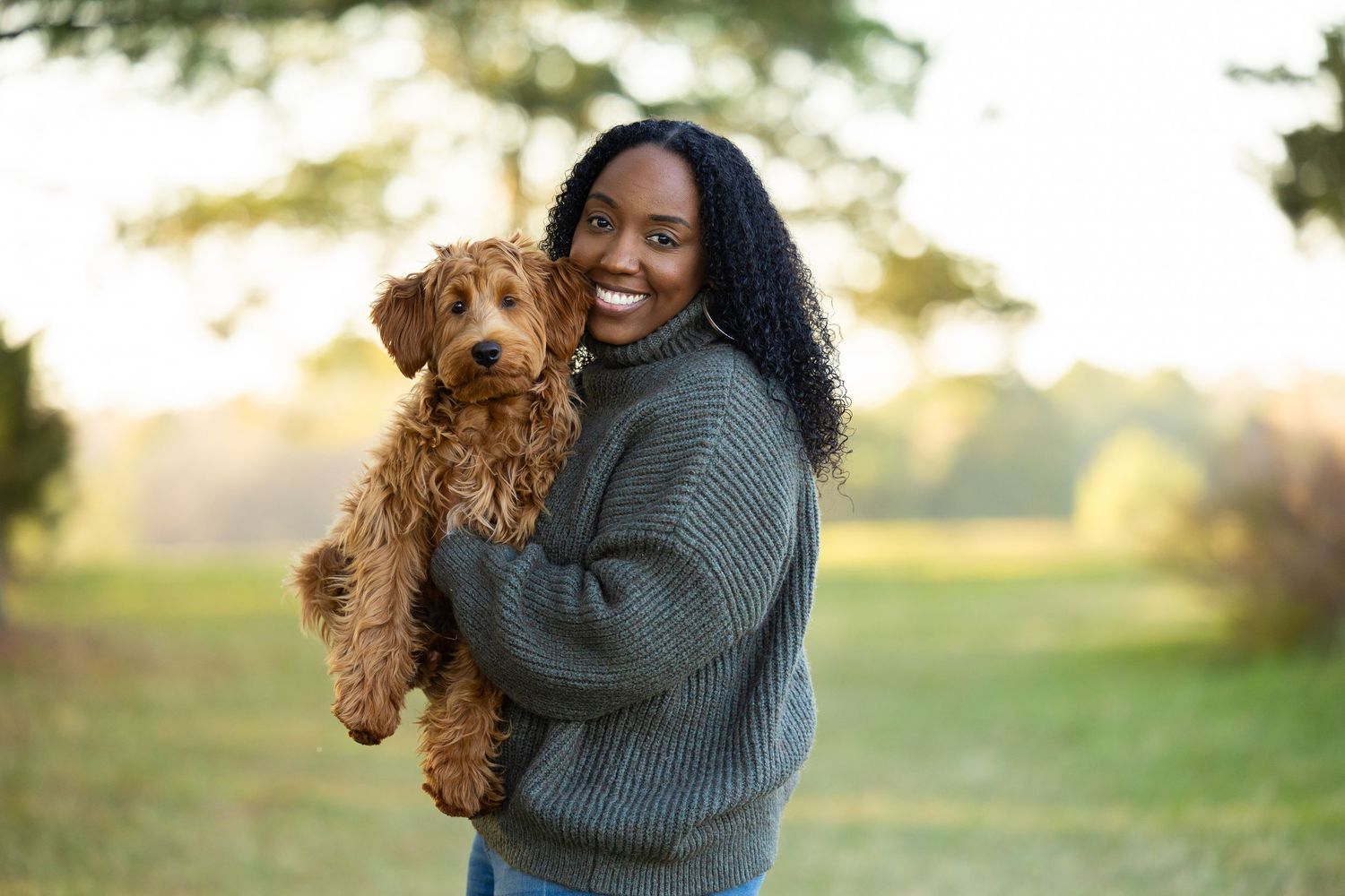 A person in a gray sweater holds a happy brown Goldendoodle puppy outdoors during golden hour.