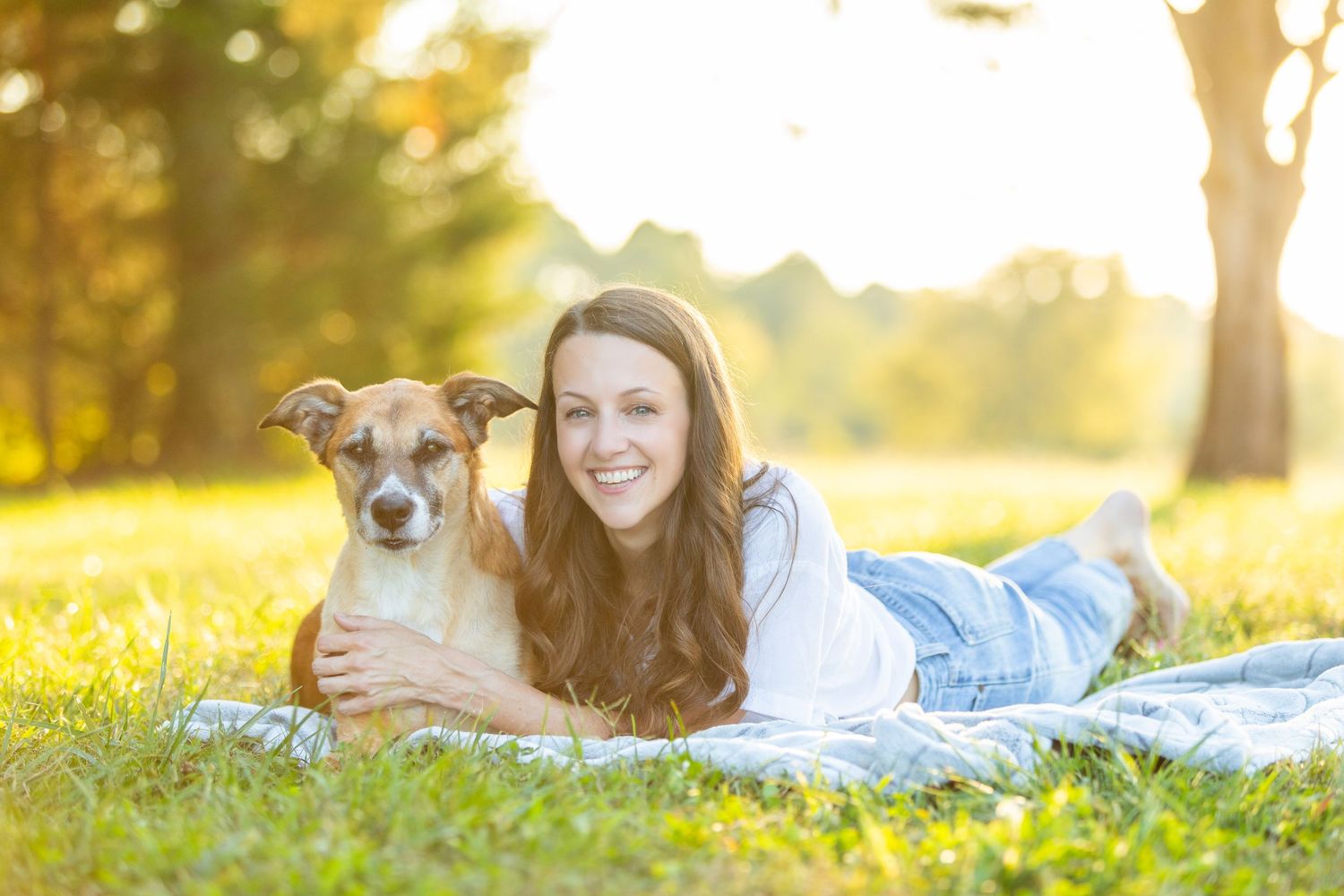 A brown and white dog and someone lying together on a blanket in a sunlit park during golden hour.