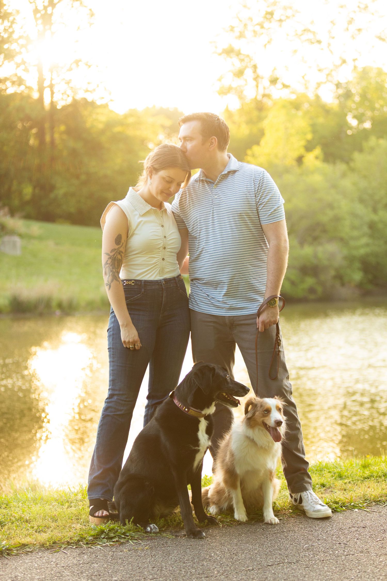 A couple sits with their Australian Shepherd dog by a serene lake at golden hour, creating a heartwarming silhouette.