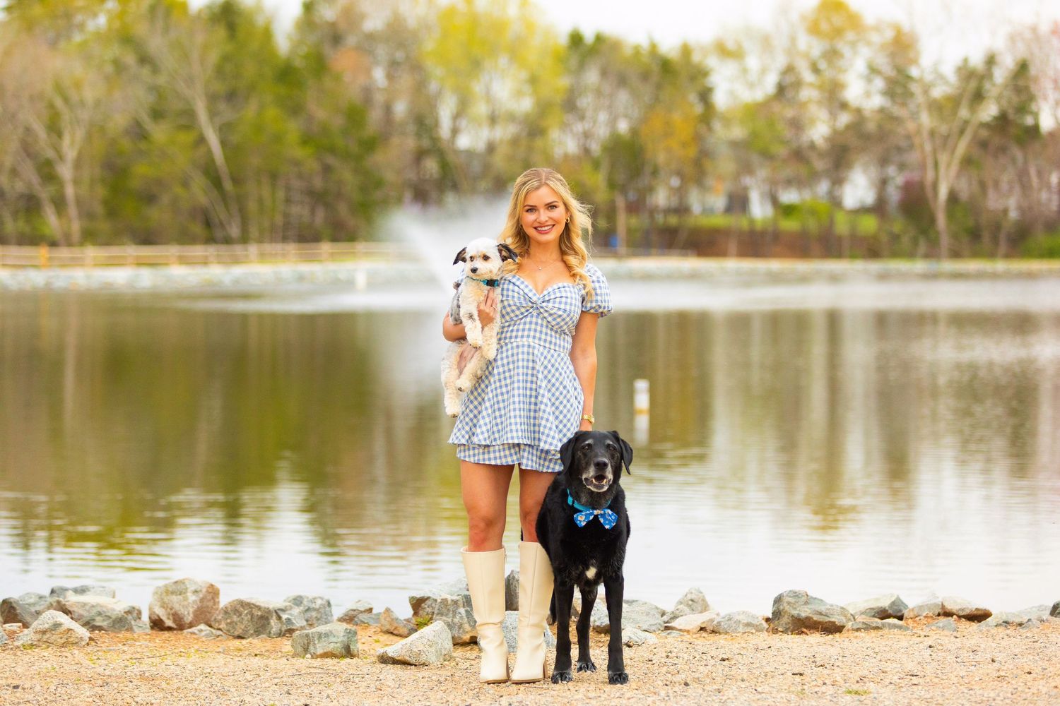 Young woman in blue dress stands by lakeside with black labrador retriever on sunny afternoon.
