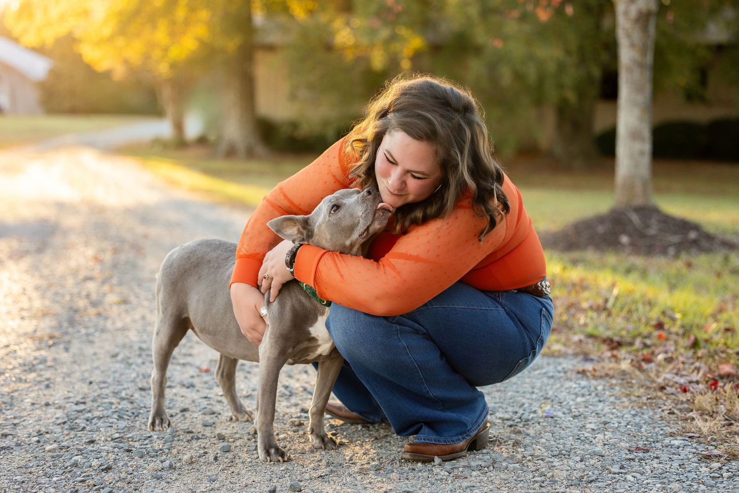 A person in blue jeans and orange jacket kneels down to pet a small gray dog on a gravel path during autumn.