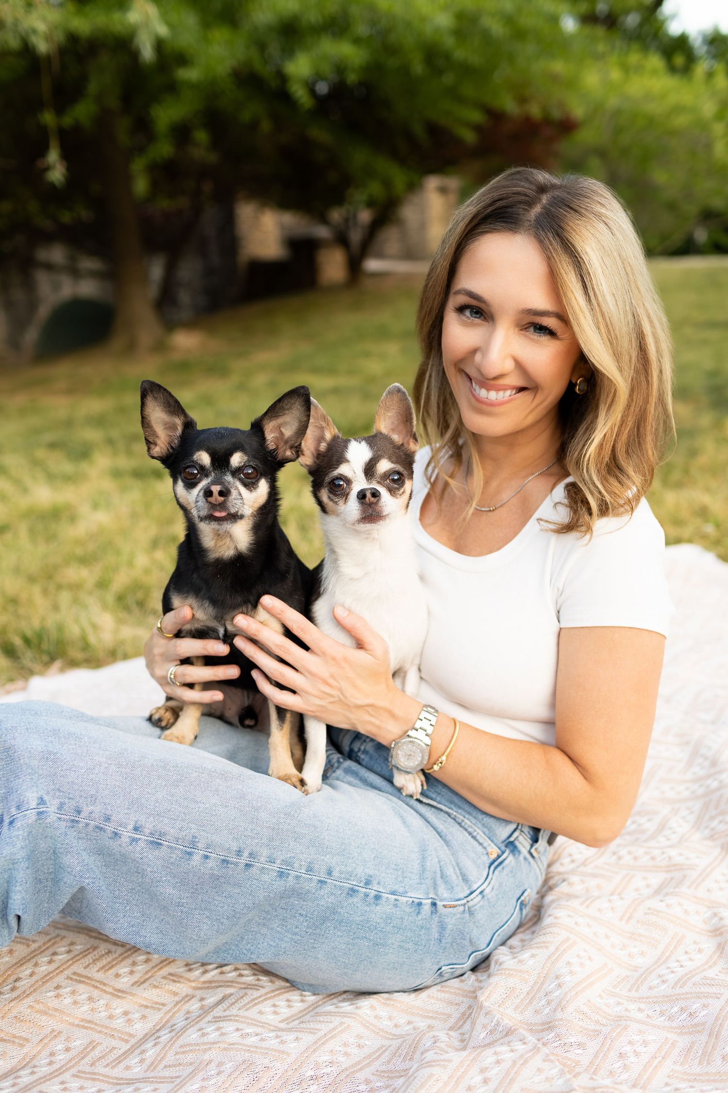 Smiling blonde woman sitting on a blanket outdoors holding two Chihuahuas in her lap.