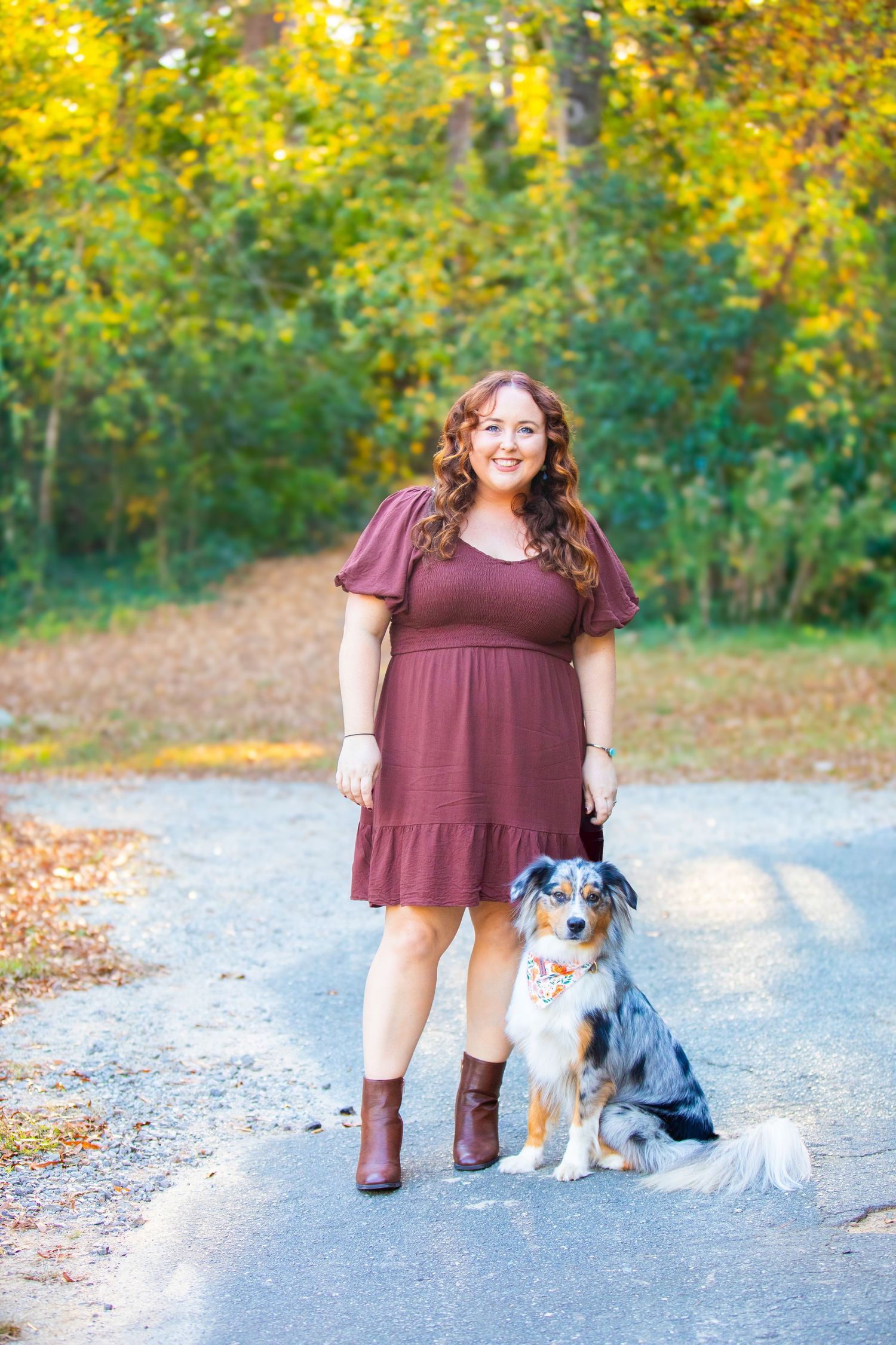 An Australian Shepherd dog sits on a path next to someone in a burgundy dress and boots during autumn.