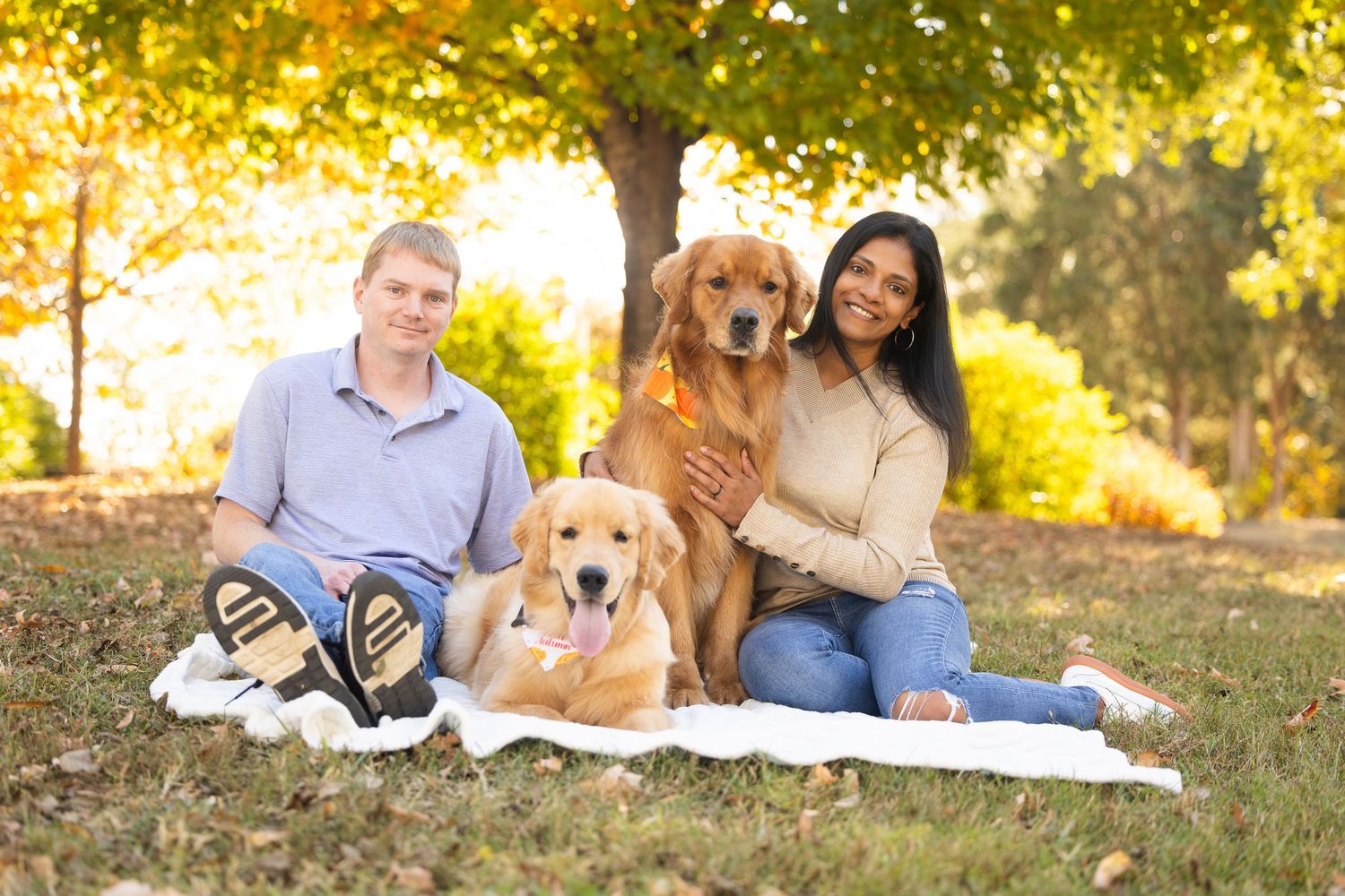 A couple sits with their golden retriever dog in a sunlit autumn park during a casual outdoor photo session.