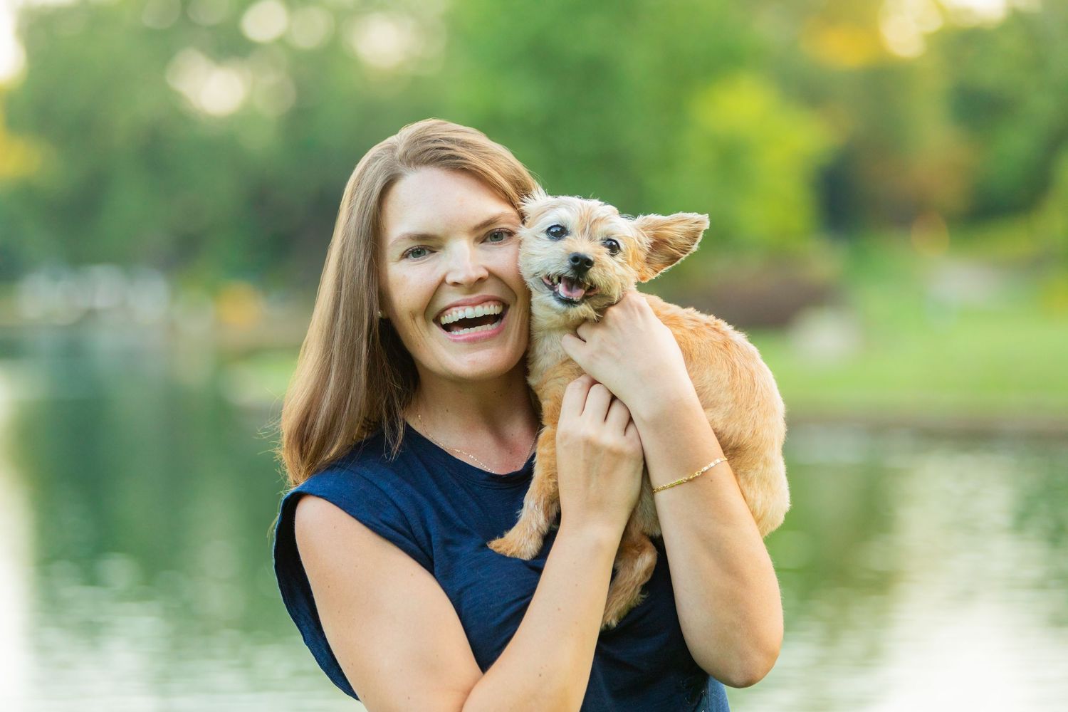A smiling woman holds a small tan Chihuahua mix near a serene park pond with lush green trees in the background.