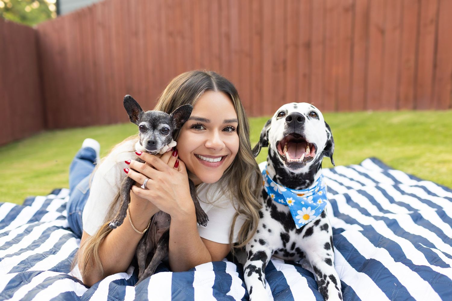 Young woman smiling outdoors on striped blanket with small Chihuahua and Dalmatian wearing blue bandana.