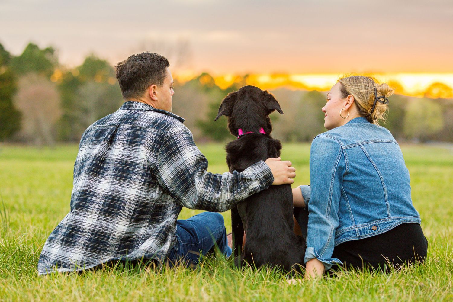 Two people sit with their black dog on grass while watching a vibrant sunset over a rural field.