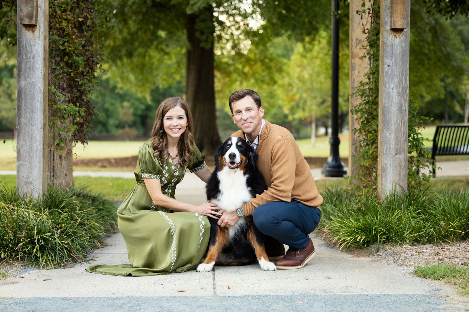 A smiling woman in an olive green dress and a man in a tan sweater crouch with their Bernese Mountain Dog on a park path framed by trees.