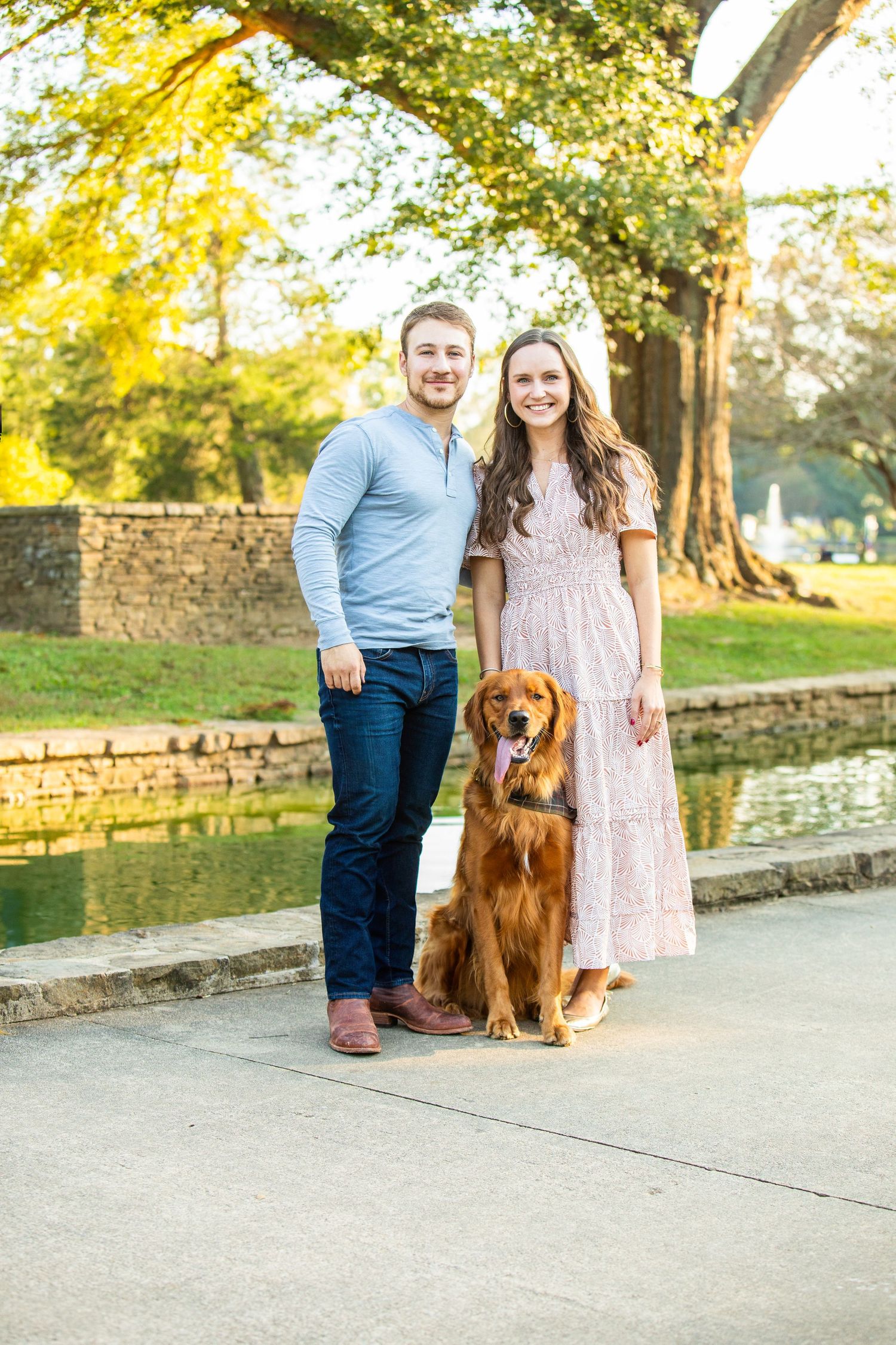 A couple stands with their Golden Retriever dog on a path near a stone wall and pond during a sunny autumn afternoon.