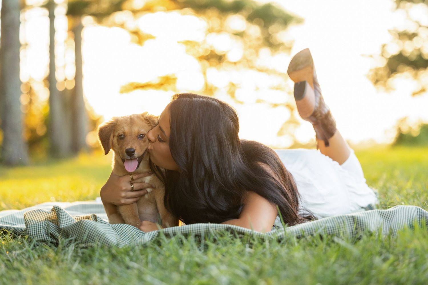 Woman lying on grass kissing her adorable golden puppy during a sunny day in the park.