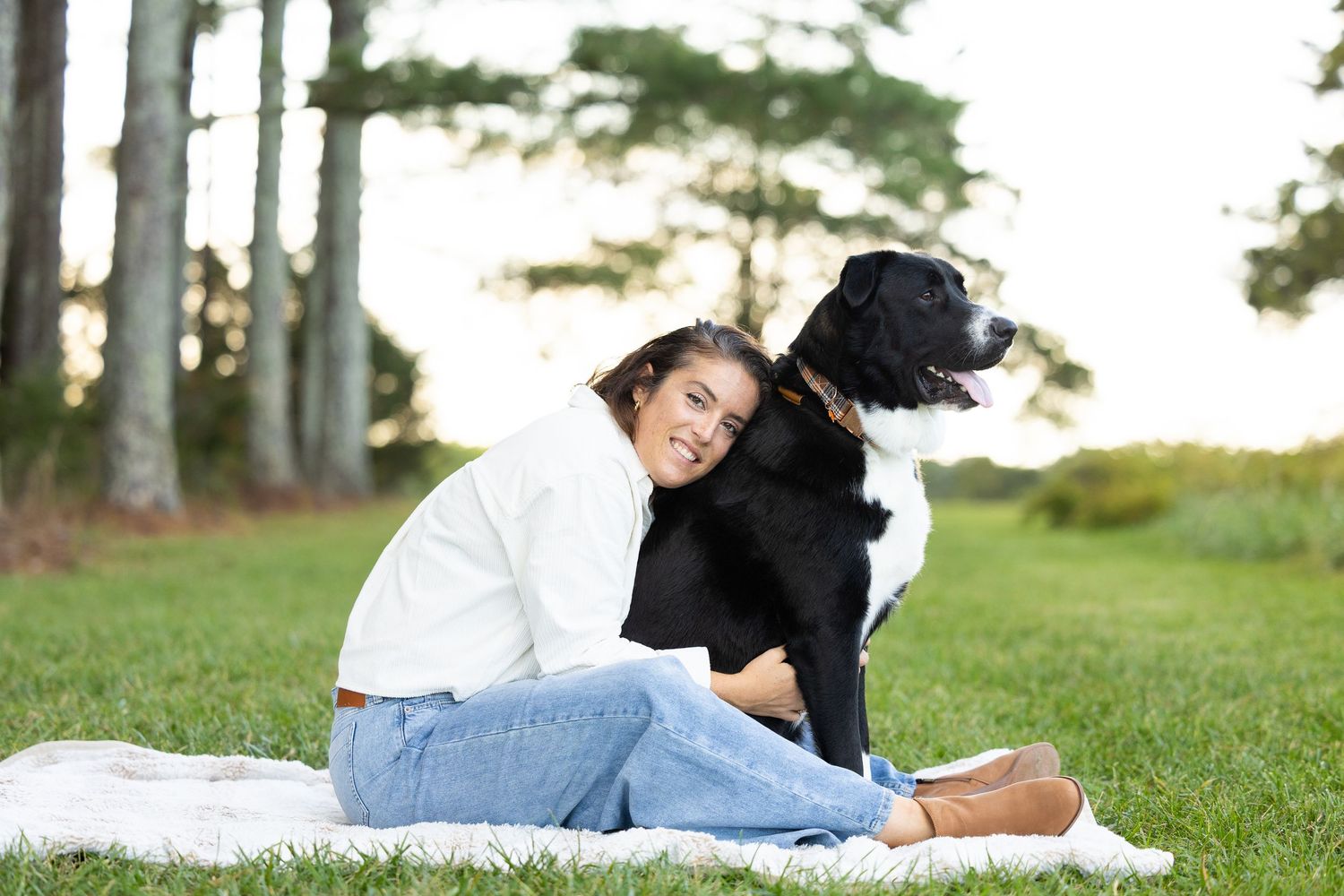 A person and black and white dog sitting together on a blanket in a grassy area with trees in the background.