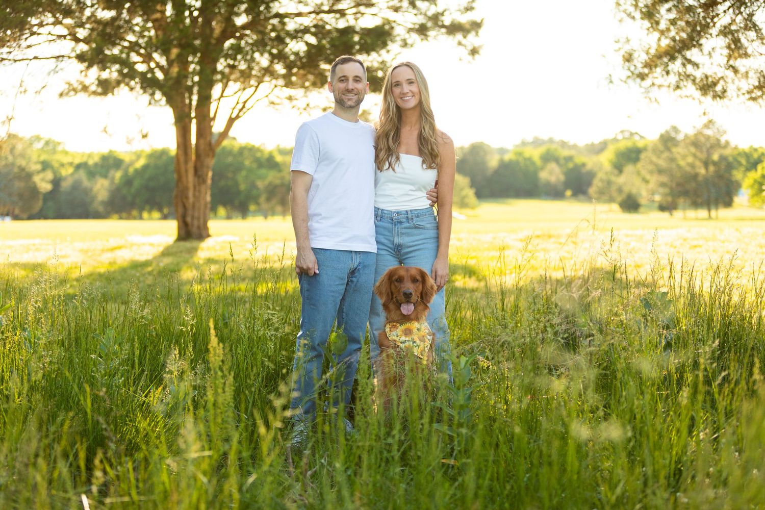 Couple and their brown dog standing in a sunny meadow with tall grass and trees at golden hour.