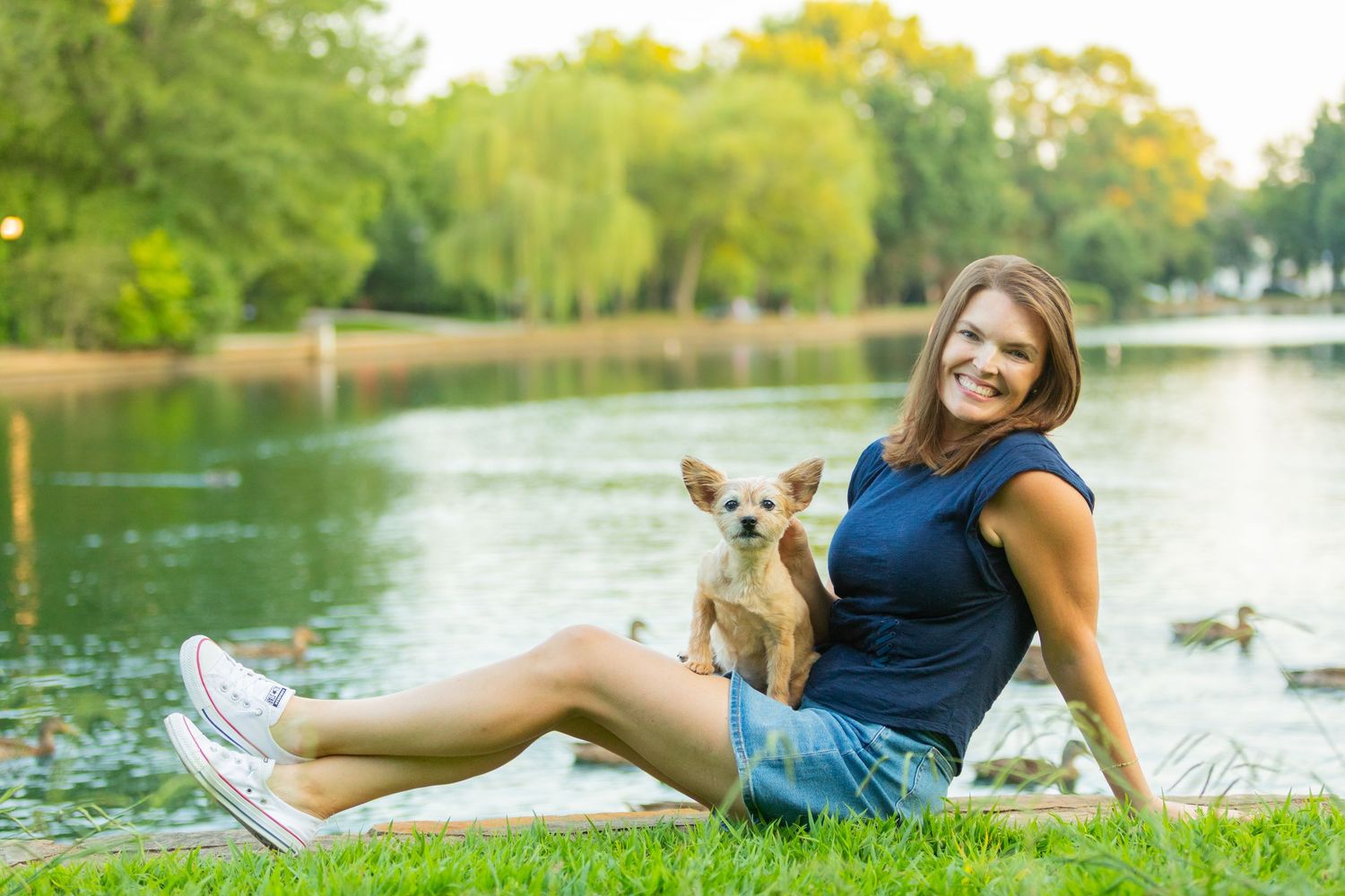 Woman smiling with Chihuahua by a serene park lake on green grass, wearing denim shorts and blue top.