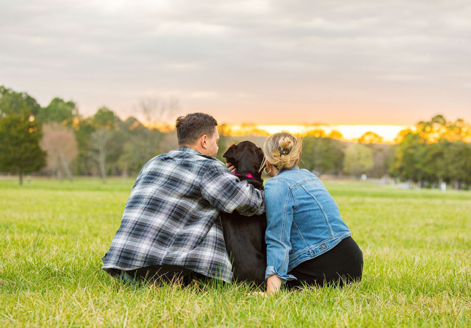 Two people in plaid shirts lie together on green grass watching a scenic sunset in the countryside.