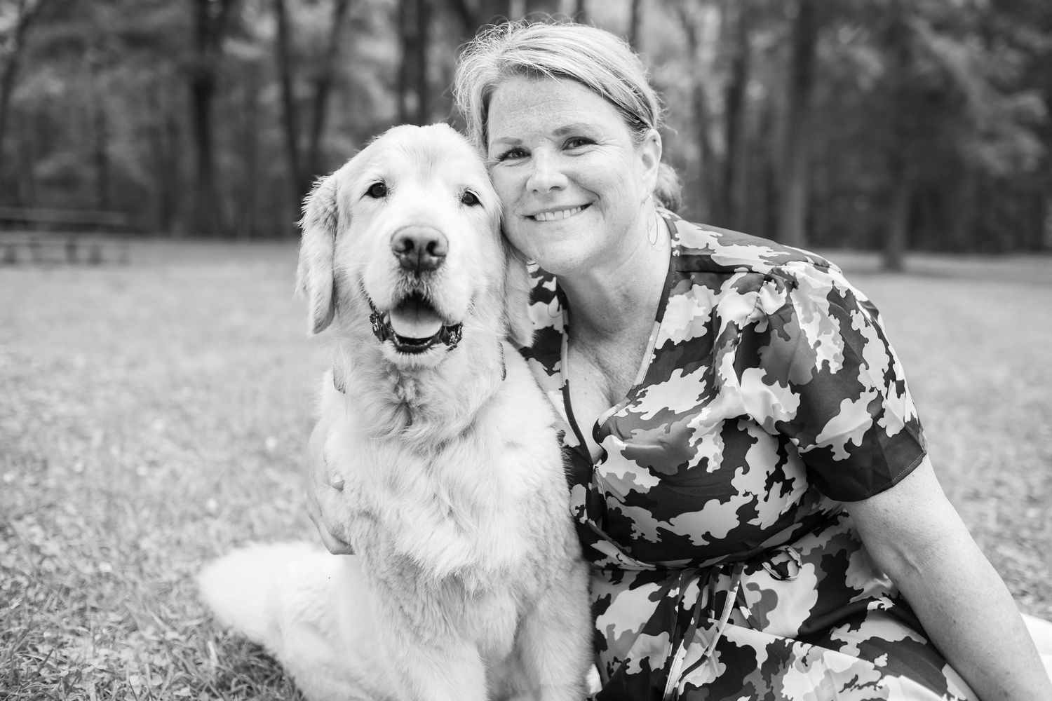 Black and white photo of a person in floral dress sitting outdoors with a happy Golden Retriever dog in grassy area.
