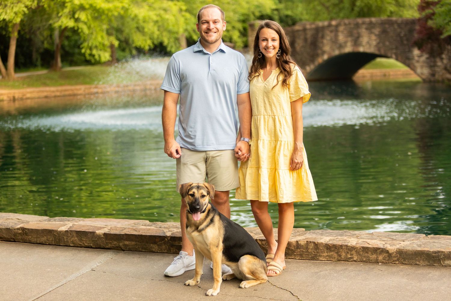 Couple walking their golden retriever dog near a serene pond with stone bridge in a scenic park setting.