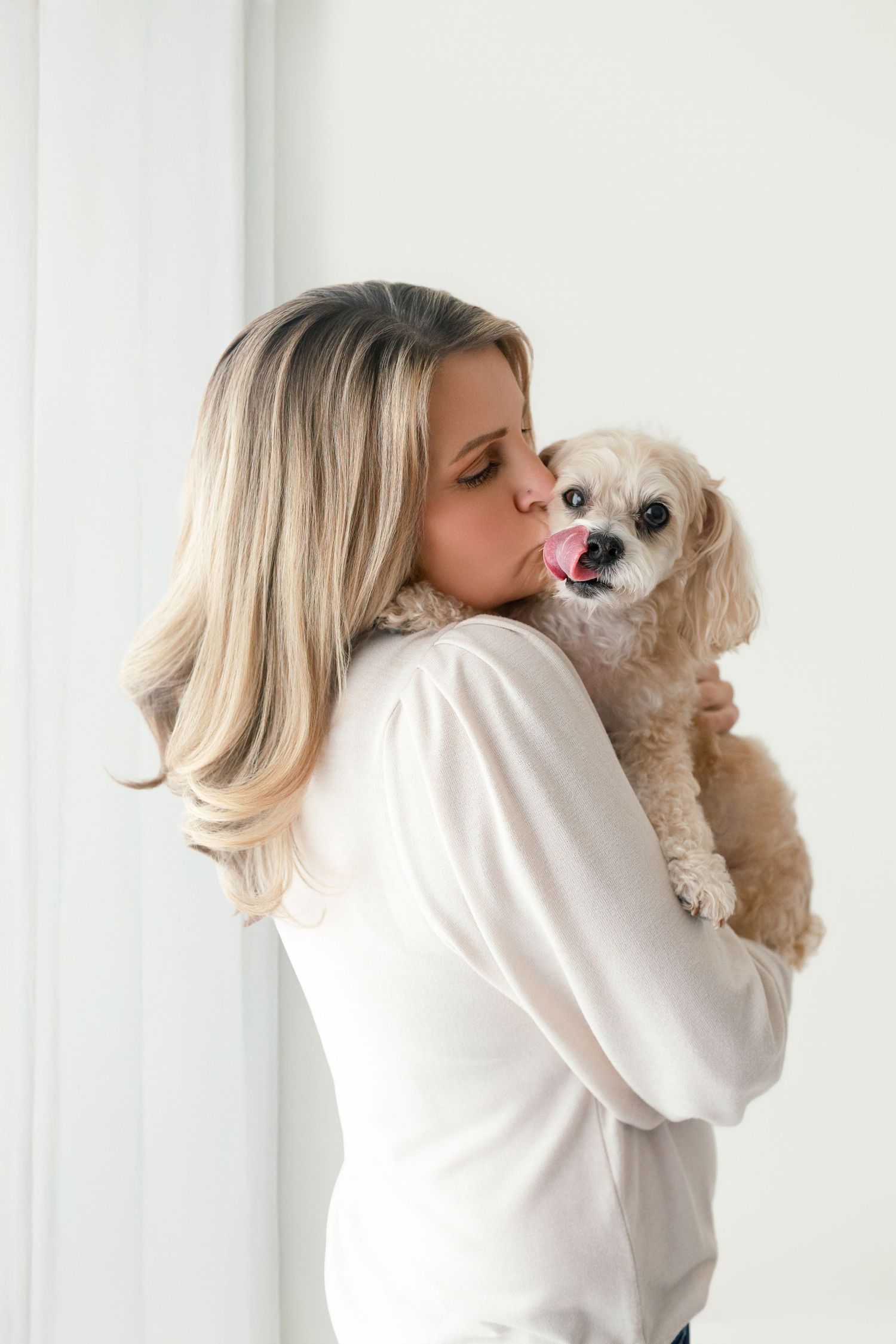 Woman in white top kissing her cute fluffy Cavapoo dog while holding it up against a light background.