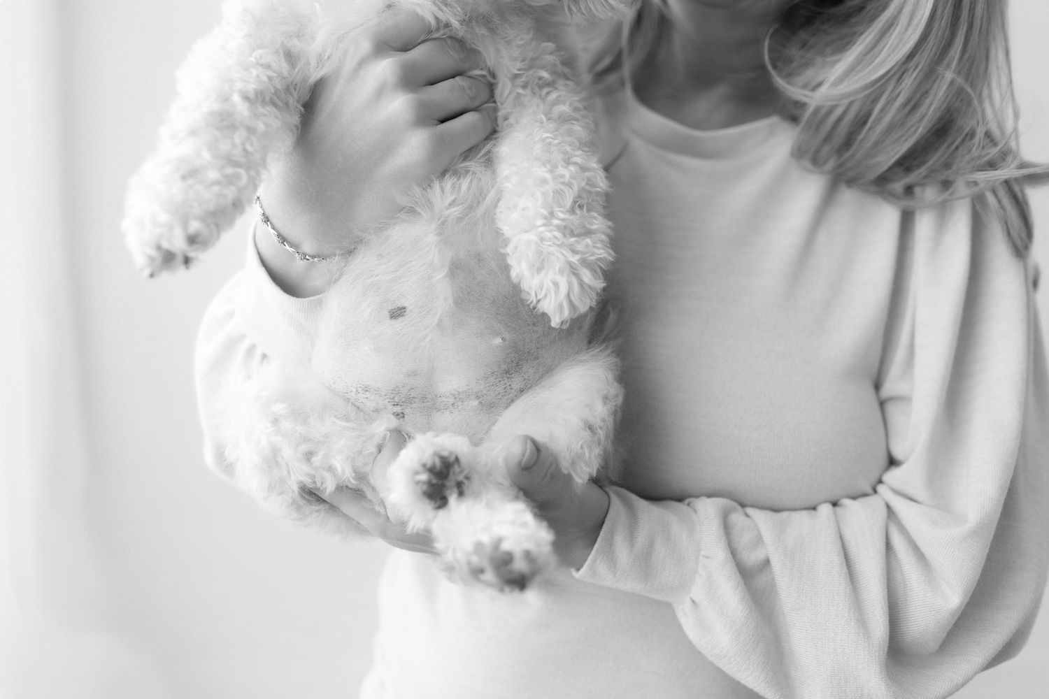 Young girl in white sweater lovingly holding a fluffy white poodle puppy in a soft black and white photo.