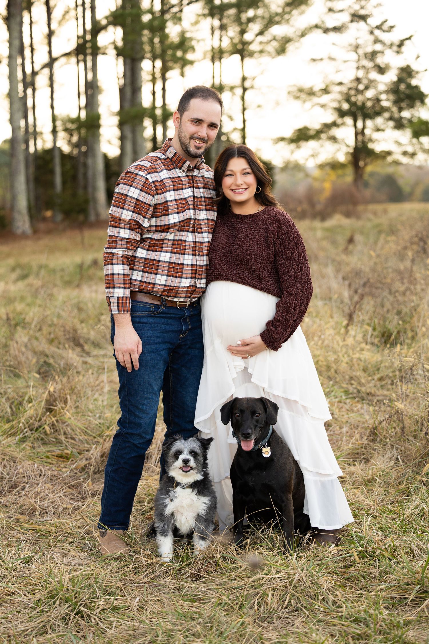 A couple poses outdoors with their two dogs - a black lab and a border collie mix - in a wooded autumn setting.