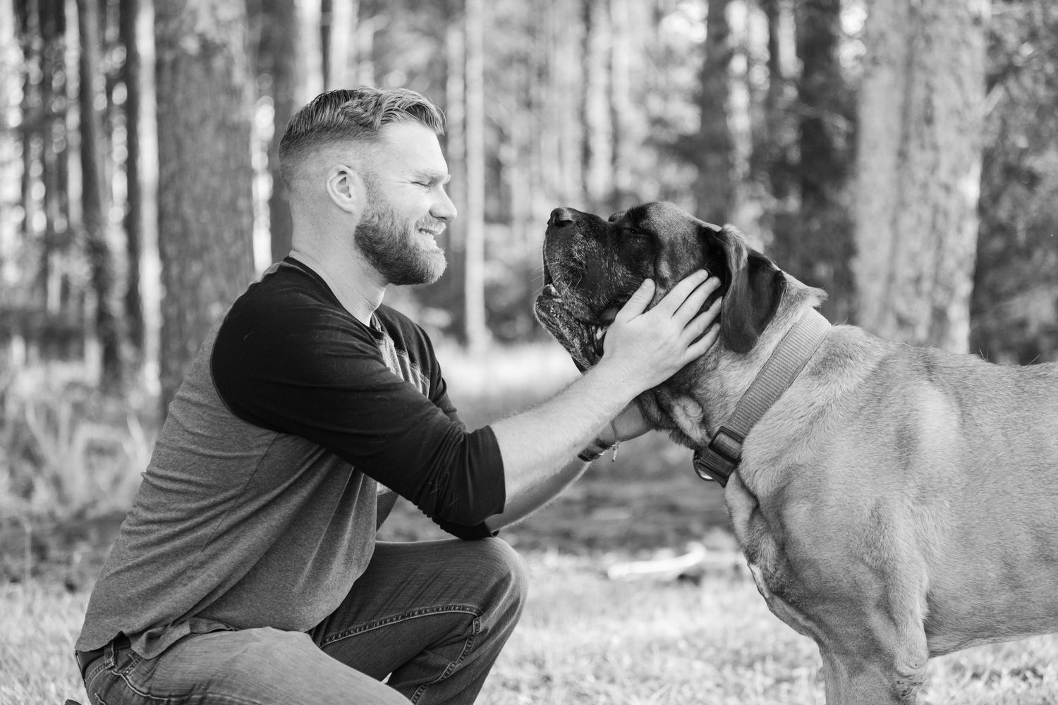 Man in black shirt sharing tender moment with French Bulldog in black and white forest setting.