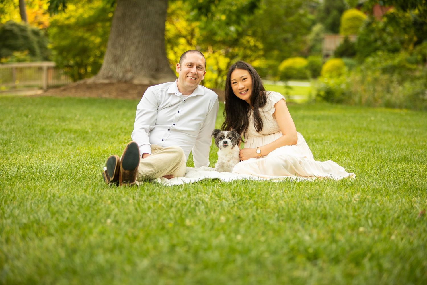 A couple lies on green grass with their fluffy white dog enjoying a sunny day in a park.