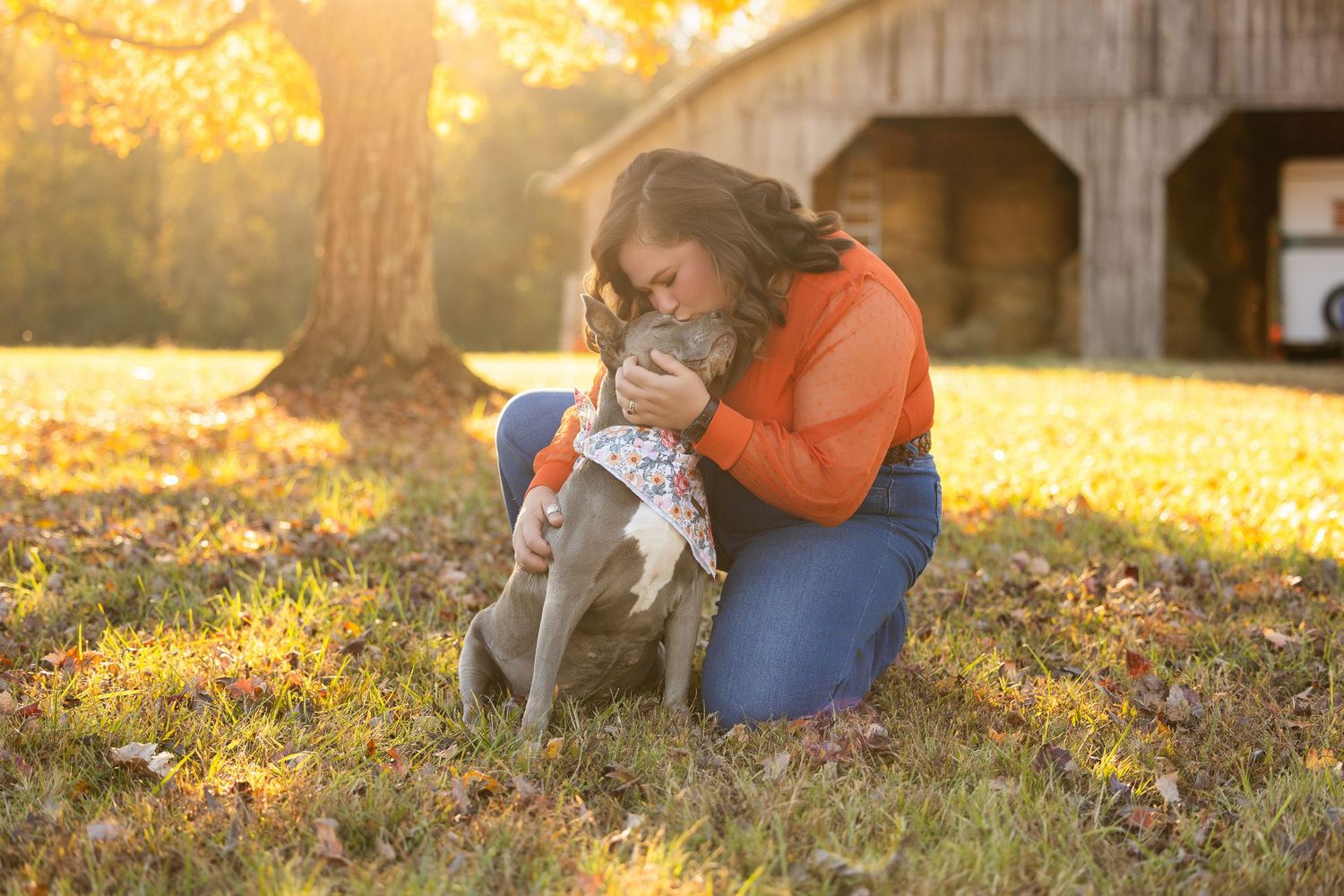 A person in orange sweater sits in autumn leaves with a dog during golden sunset light.