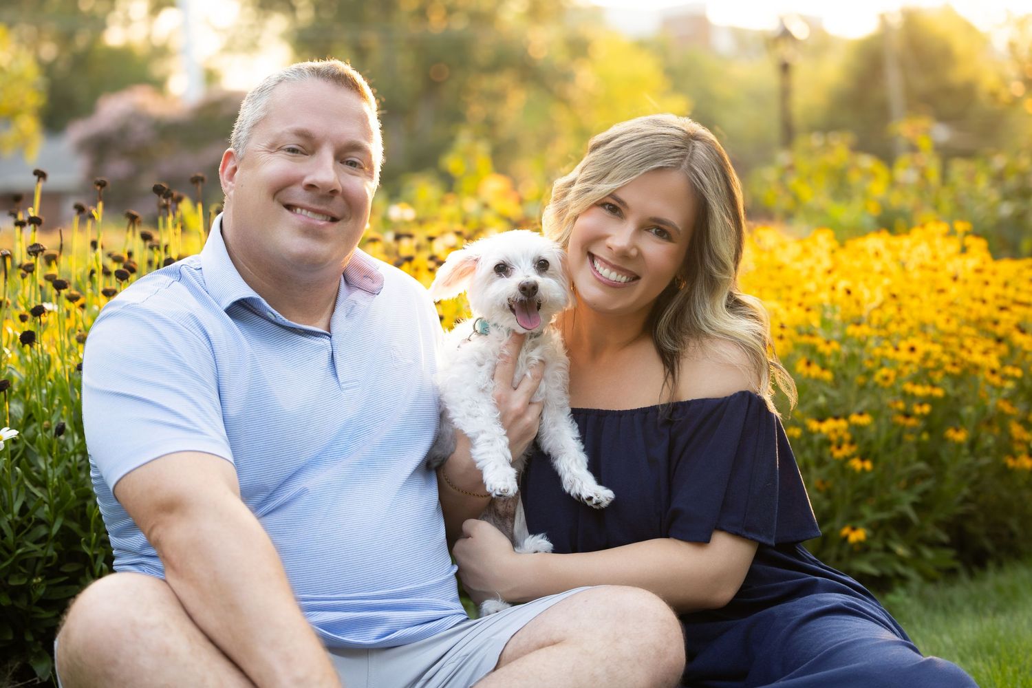 A smiling couple sits outdoors holding a small white dog, surrounded by vibrant yellow flowers in a sunny garden.