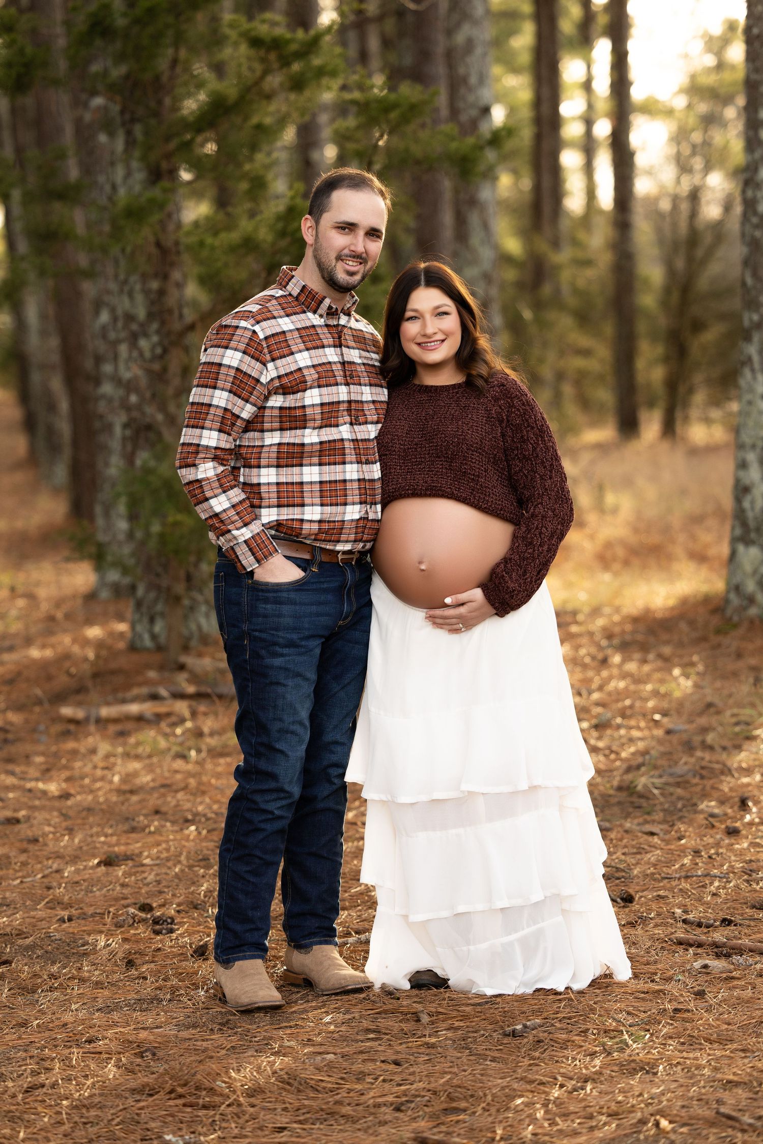A couple in casual attire poses for maternity photos in a sunlit forest setting during autumn.