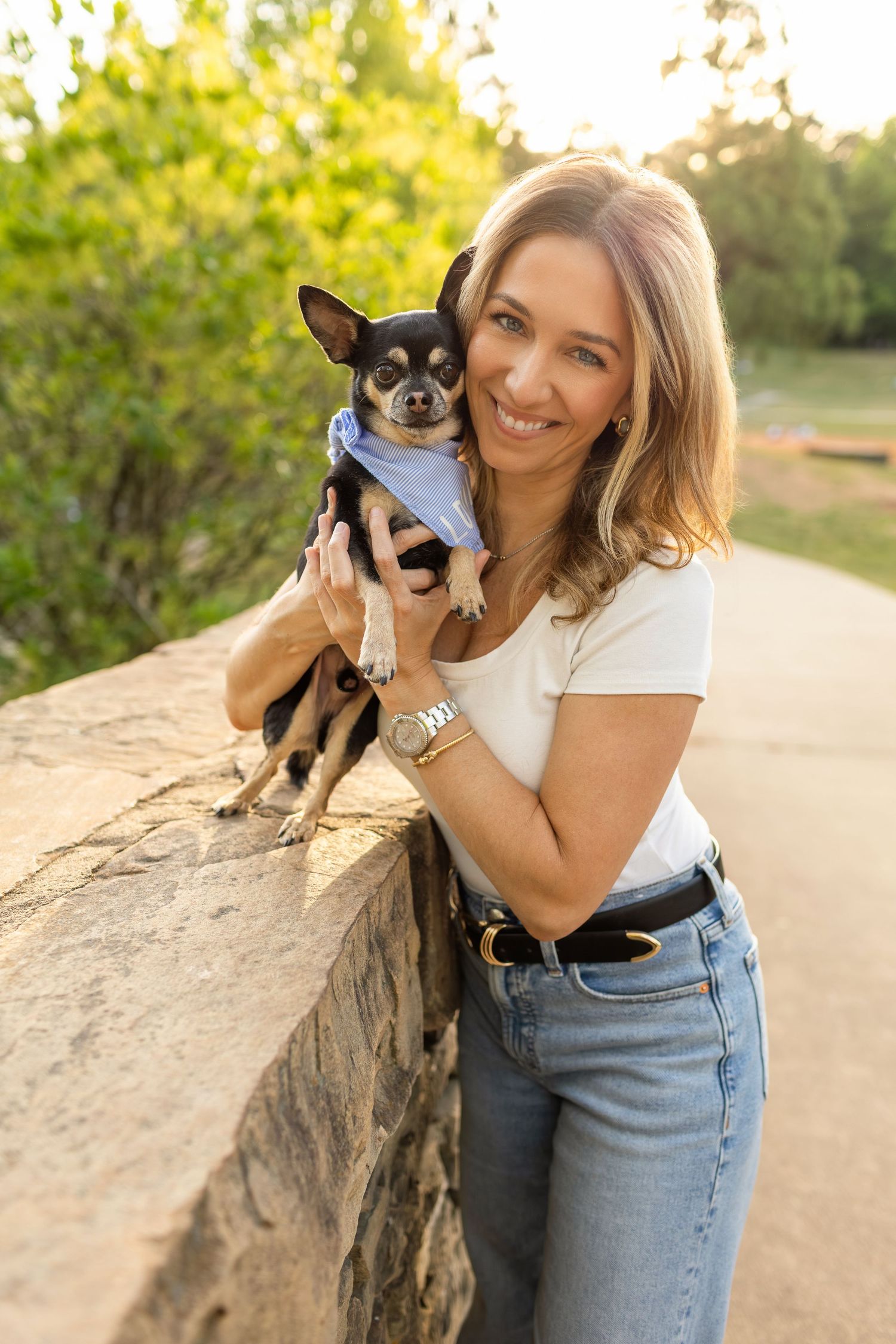 Smiling blonde woman holding a small black Chihuahua wearing a bandana outdoors in golden hour light.