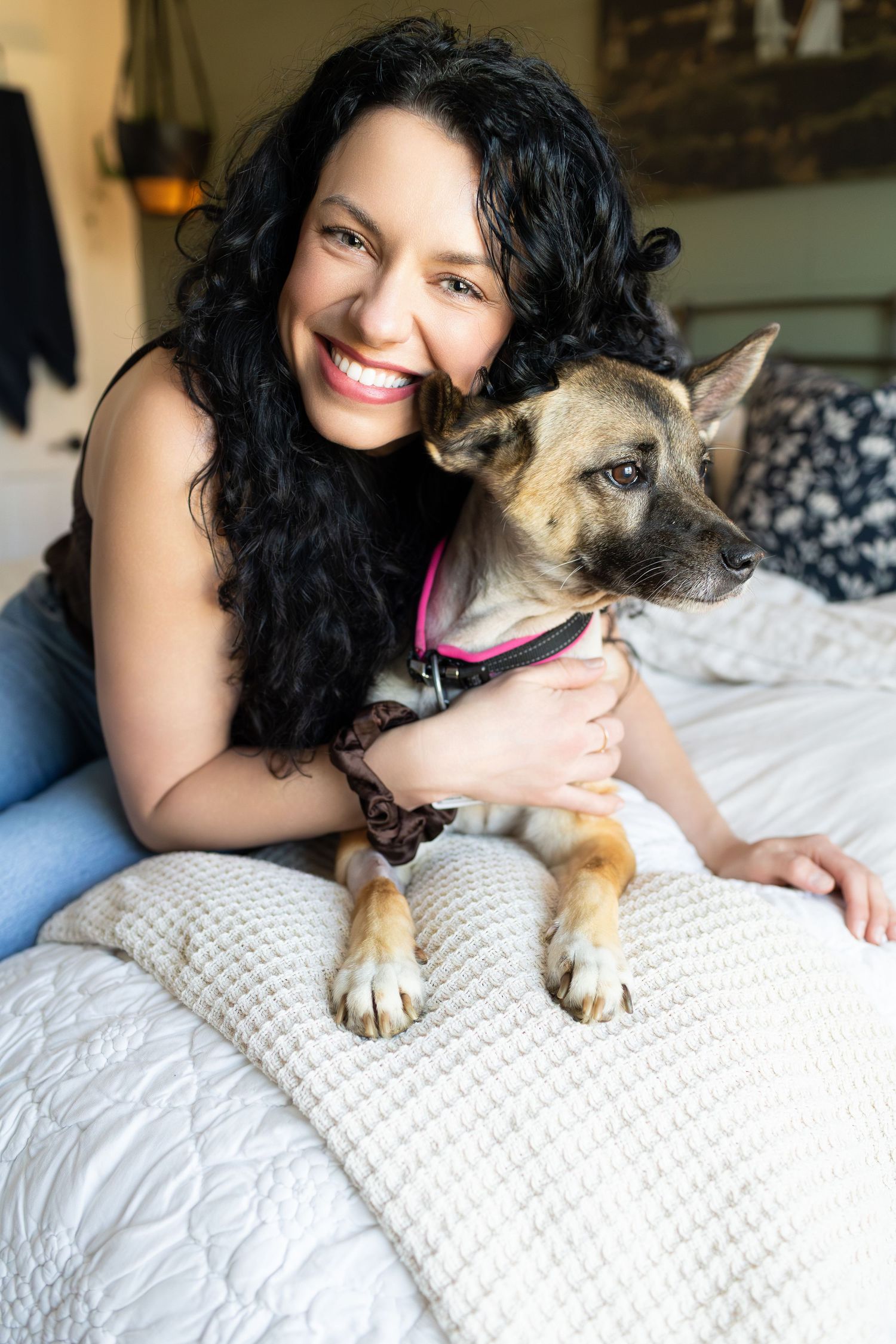 Smiling woman with curly black hair hugging her tan dog on a white bed in a cozy bedroom.