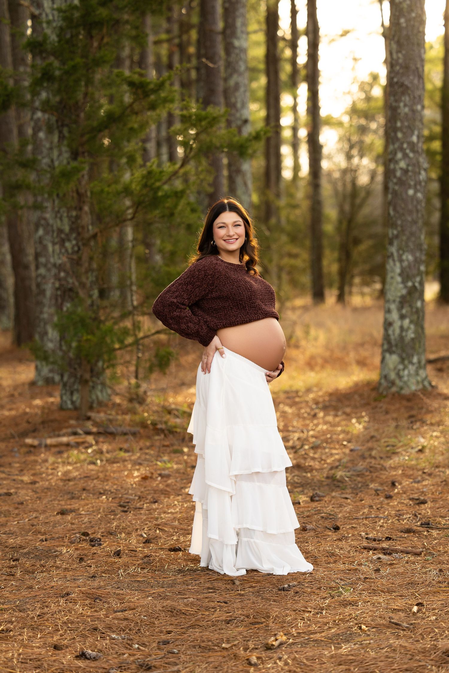 A maternity photo in a forest setting shows an expectant mother wearing a brown sweater and flowing white tiered skirt.