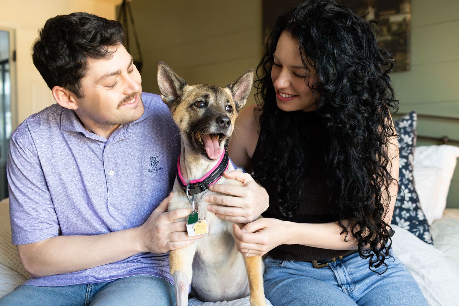A happy couple smiling and holding an excited German Shepherd puppy with a pink collar indoors.