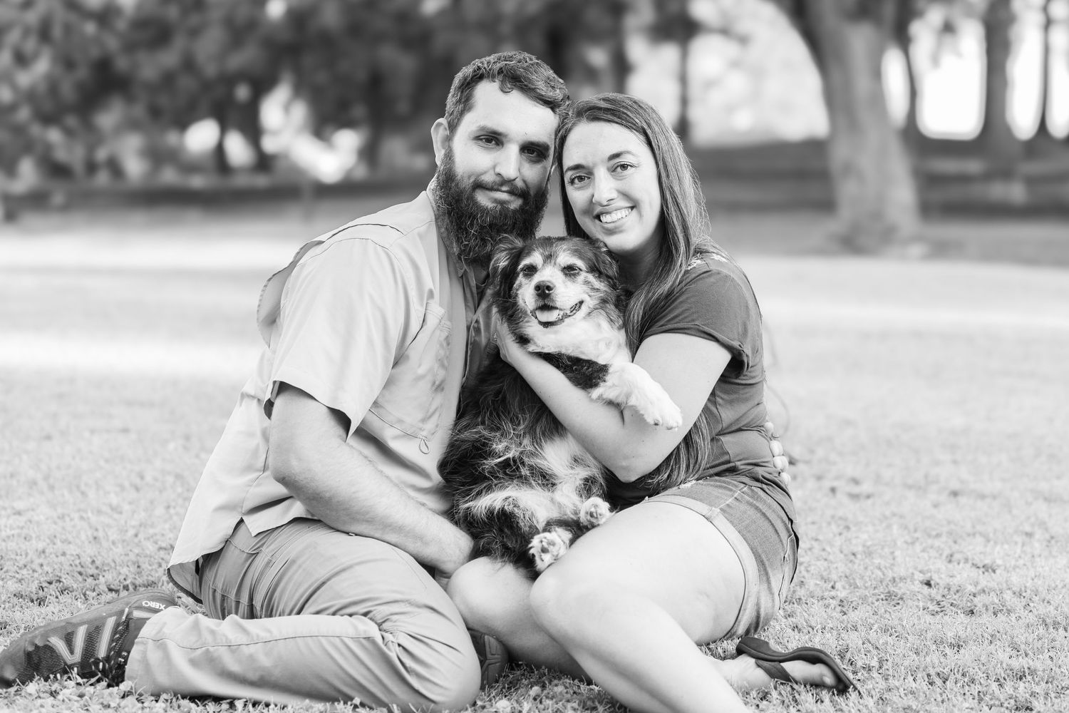 A couple sits in a grassy park with their fluffy dog while posing for a black and white portrait.