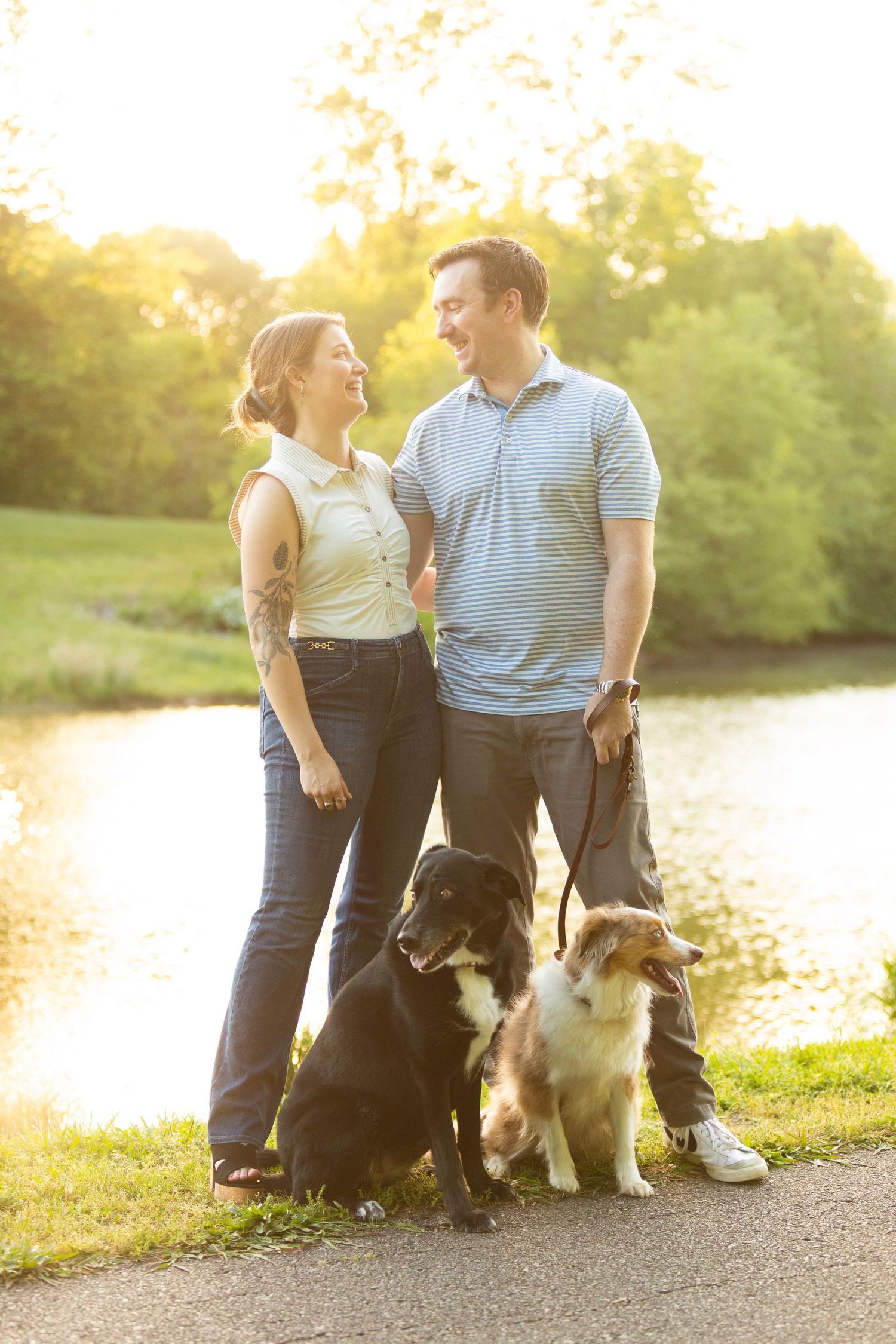 Two people sit with their dogs on a path by a lake at sunset in a golden-lit outdoor setting.