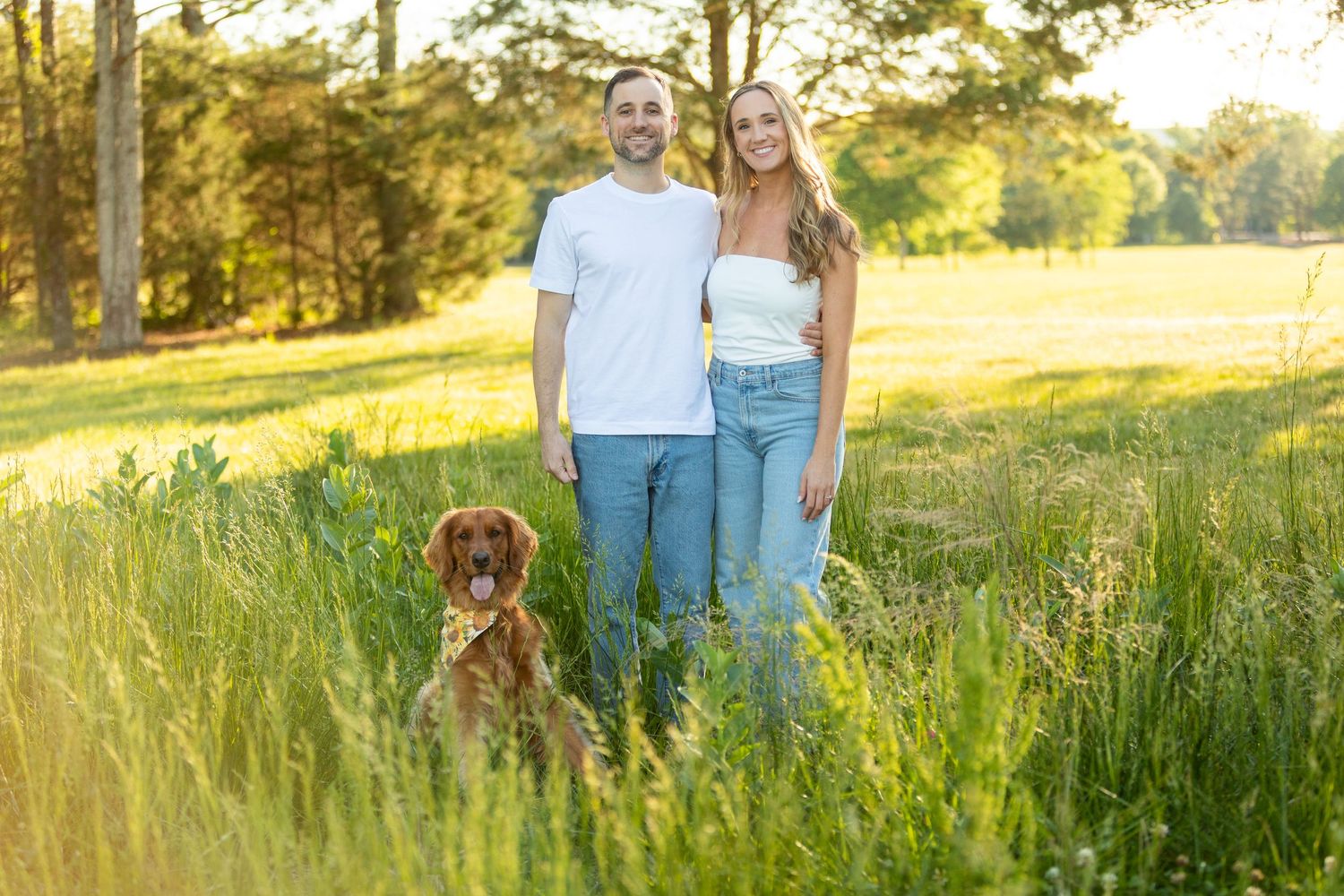 A couple and their golden retriever dog pose together in a sunlit grassy field during golden hour.
