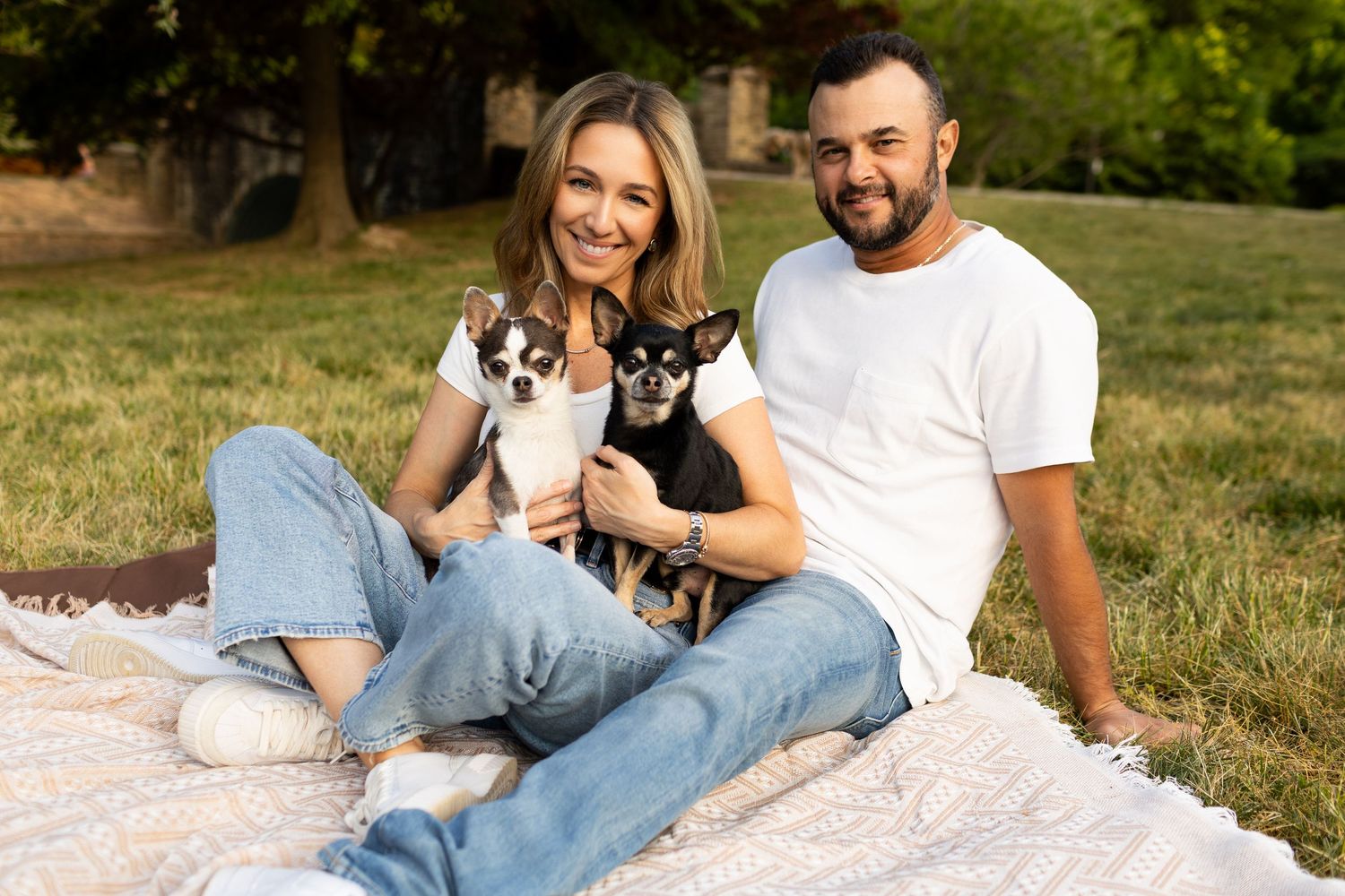 Happy couple sitting on blanket in park holding two small Chihuahua dogs, smiling outdoors on sunny day.