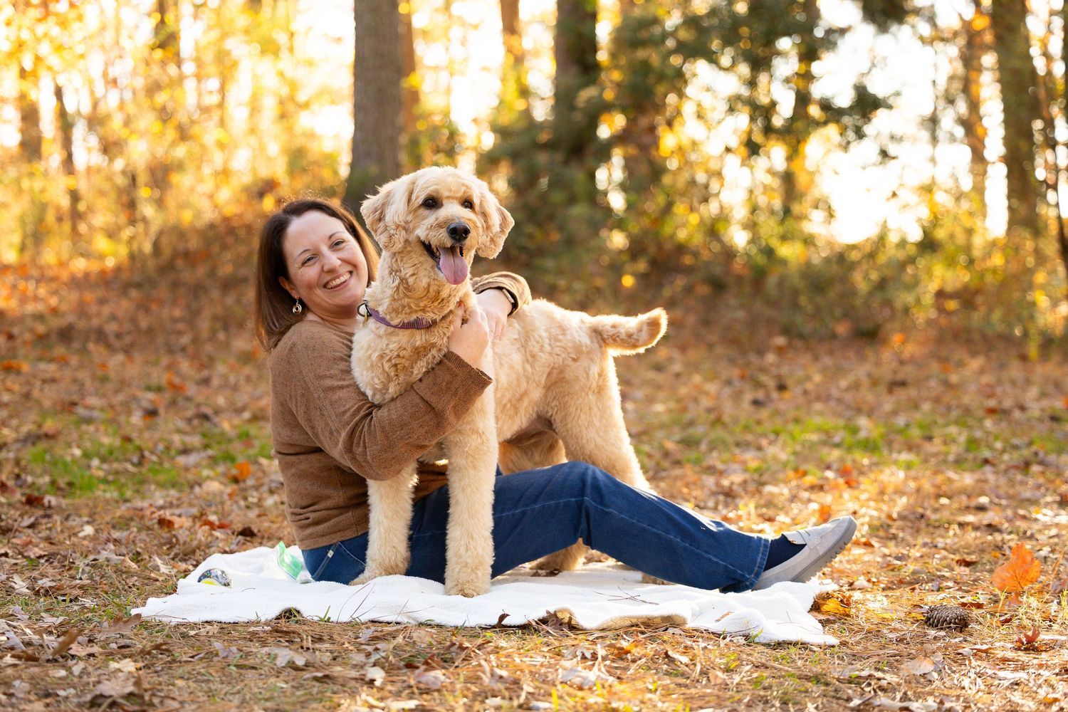 Woman smiling and hugging a fluffy Goldendoodle dog outdoors in an autumn forest setting.