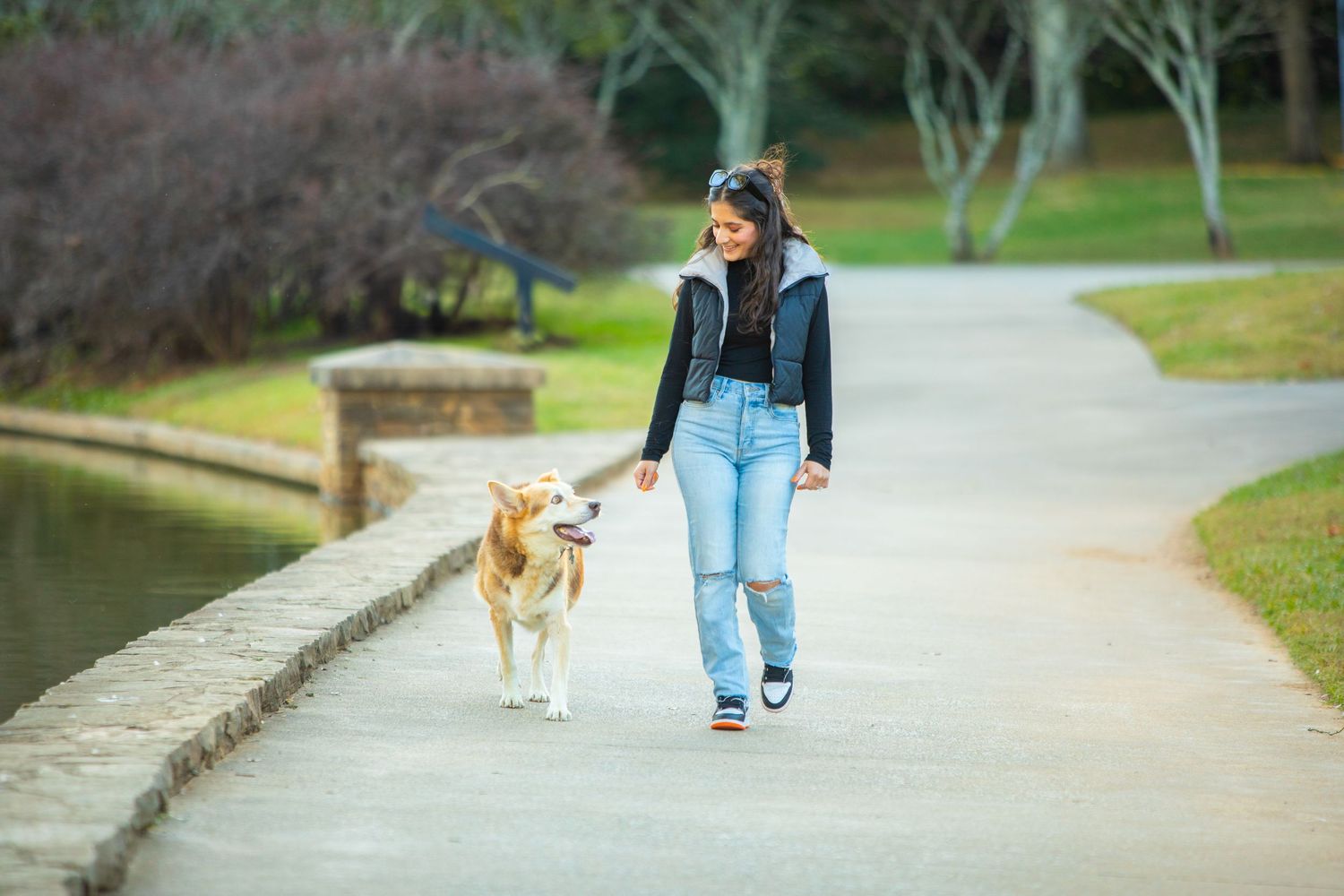 A person in blue jeans and dark jacket walking a light-colored dog on a paved path beside a pond in a park.