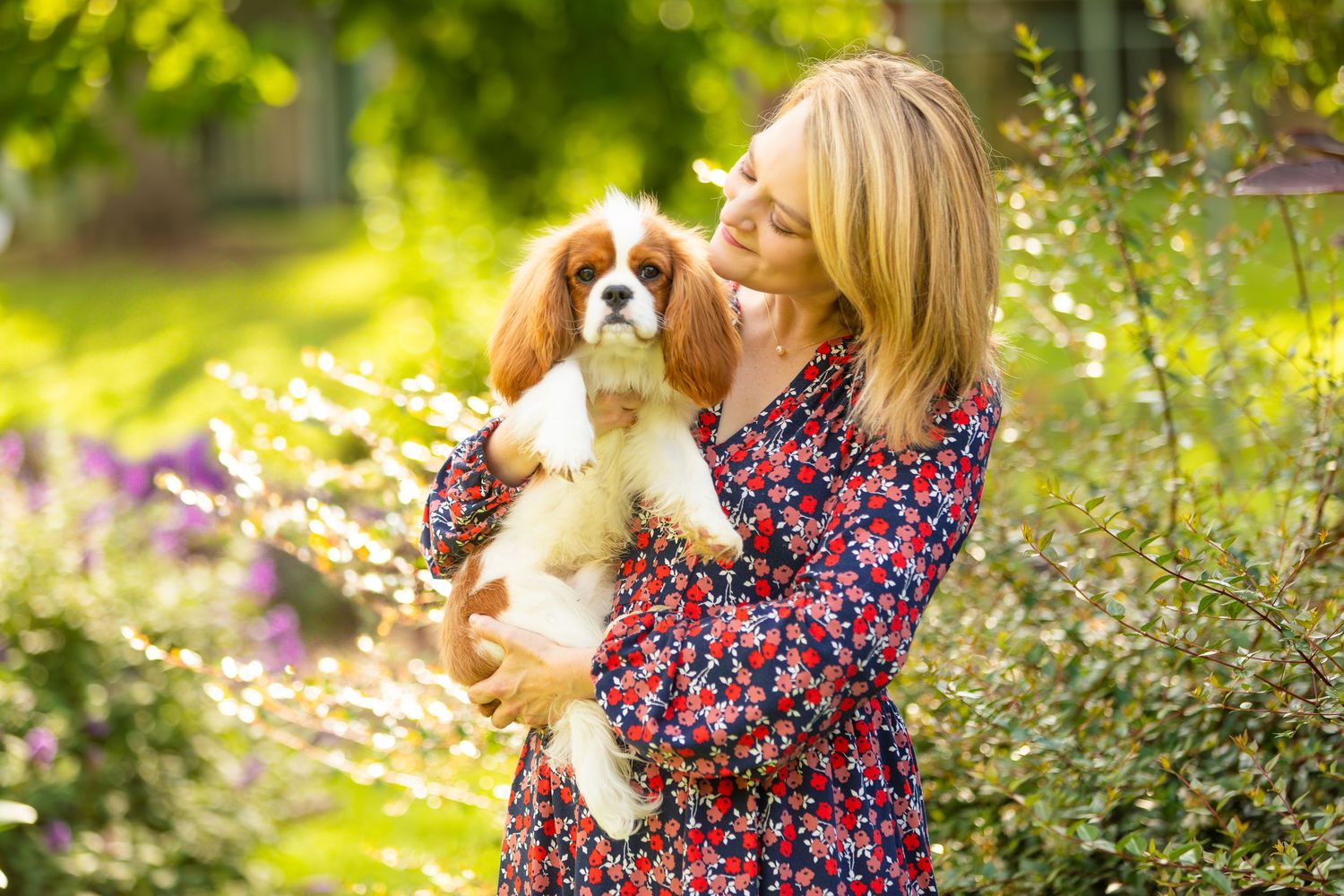 A Cavalier King Charles Spaniel puppy is held lovingly outdoors in a garden with blooming flowers and sunlit greenery.