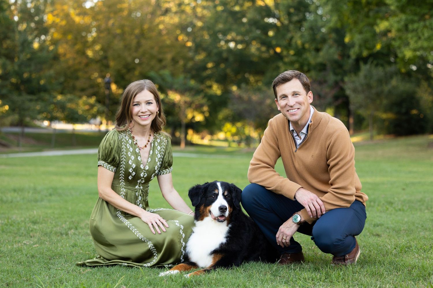 A smiling woman in a green embroidered dress and a man in an orange sweater pose outdoors on grass with a Bernese Mountain Dog.