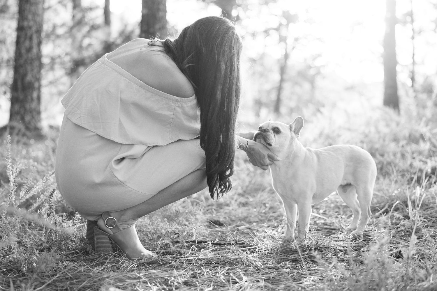 Black and white photo of a person crouching down to interact with a small white dog in a forest setting.
