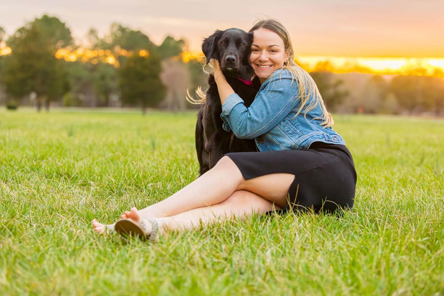 A black Labrador and companion embrace in a grassy field at sunset during a heartwarming outdoor photo session.