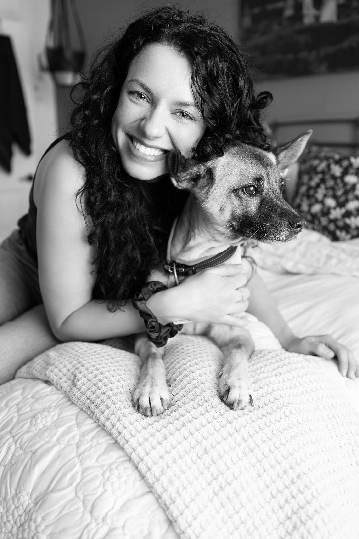 Black and white photo of a person with long dark curly hair cuddling with their German Shepherd dog on a textured bedspread.
