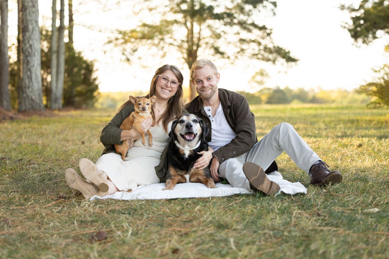 A couple sits on a blanket in a grassy field at sunset with their black and white dog.
